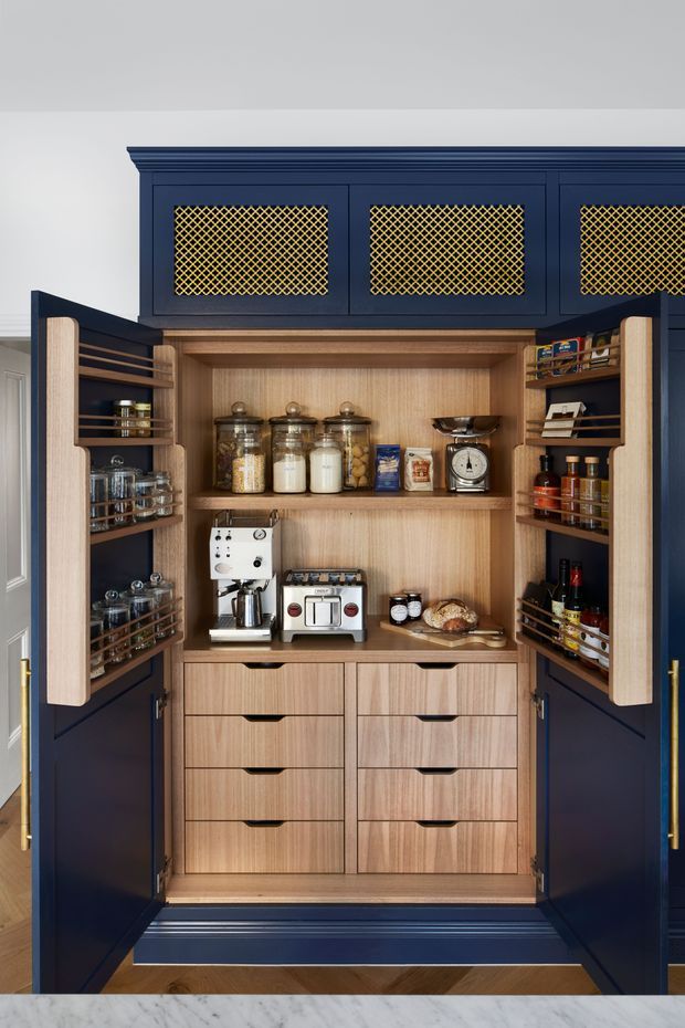 A detailed built-in pantry cupboard shown open, with dark navy painted cabinetry, warm timber interior joinery, drawers, shelves, and door-mounted racks. The pantry is stocked with jars, bottles, dry goods, coffee equipment, scales, and bread. Decorative gold lattice panels occupy the upper cabinet doors, with brass handles and traditional moulded trim.