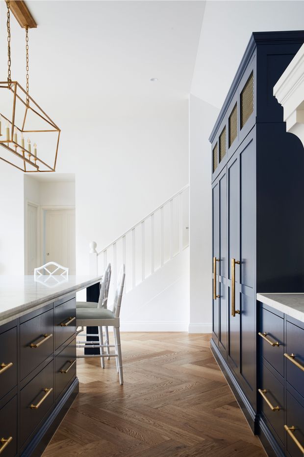 Bright, refined kitchen interior with deep navy shaker-style cabinetry, brass hardware, pale stone benchtops, and a warm herringbone timber floor. A central island with stools sits beside a white staircase and entry opening, while a geometric brass pendant adds a decorative focal point. The cabinetry features framed doors and upper mesh-front panels, creating a classic yet contemporary look.