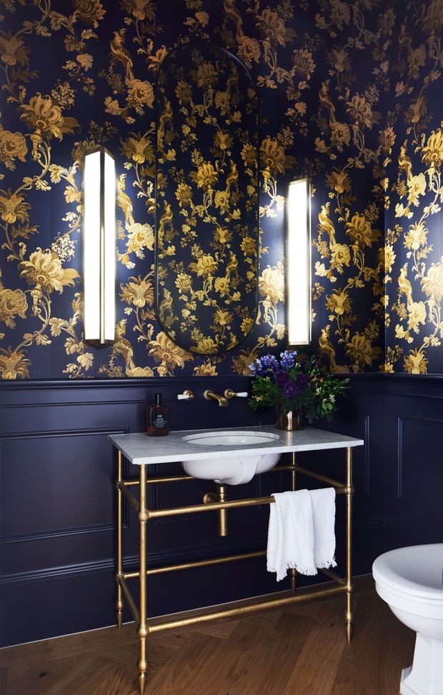 A compact, dramatically styled bathroom featuring deep navy paneled walls and ceiling covered in ornate gold botanical wallpaper. A tall arched mirror is flanked by vertical illuminated sconces above a white vessel sink set into a pale stone countertop on an exposed brass console vanity. A white towel hangs from the brass frame, with a toilet partially visible at right. The floor is warm herringbone timber, creating a luxurious, theatrical contrast with the dark walls and gilded detailing.