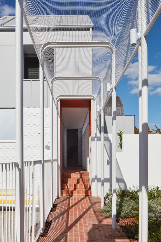 A bright exterior passageway beside a contemporary building, enclosed by a distinctive curved white mesh canopy and supported by slim white tubular frames. The walkway and steps are paved in red brick, leading toward a recessed entrance with a vivid terracotta-red ceiling and side wall. White rendered and textured masonry walls, metal railings, strong sunlight, and deep shadows create a crisp, graphic facade composition.