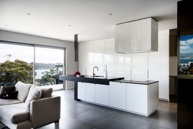 Contemporary open-plan kitchen and living area with a large white island, black stone-look benchtops, glossy white cabinetry, and a textured suspended rangehood enclosure. Floor-to-ceiling glazing provides a waterfront outlook, while a neutral sectional sofa occupies the living zone. The space is bright, refined, and minimalist in character.