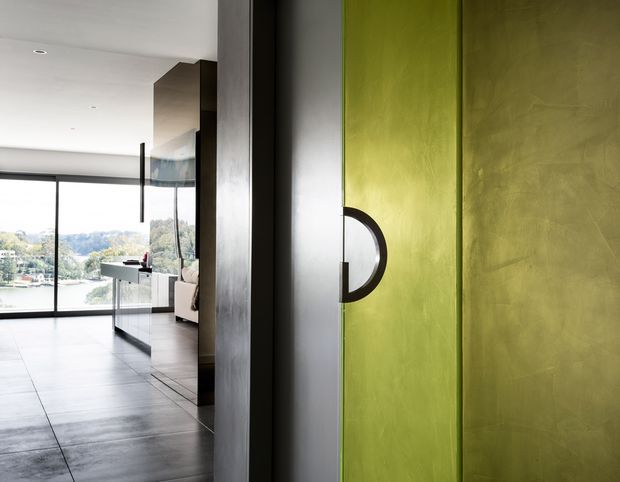 View through a modern interior toward floor-to-ceiling glazing overlooking a waterfront landscape. A vivid lime-green feature door or pivot panel with a bold circular black-and-white handle dominates the foreground, while dark tiled flooring, dark vertical joinery, and streamlined cabinetry create a sleek contemporary setting.