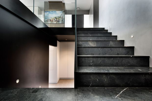 Interior view of a contemporary stairway with broad dark stone-clad treads and risers. The stairs rise beside a dark built-in wall or balustrade, with a glass guardrail and minimalist recessed wall lights. Pale cabinetry and a bright lower-level space are visible beneath and beyond the stair, while reflective glazing and a framed artwork appear at the upper landing. The palette is predominantly charcoal, black, grey, and white, creating a refined, dramatic atmosphere.