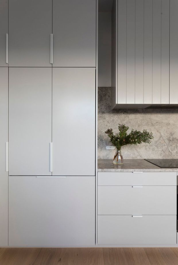 A close-up view of a sleek contemporary kitchen with tall matte pale-grey cabinetry, integrated appliance storage, long vertical handles, lower drawers, and a dark stone benchtop. The backsplash features a mottled natural-stone or concrete-like finish, with a cooktop and a small vase of greenery. Light timber flooring adds warmth to the otherwise restrained neutral palette.