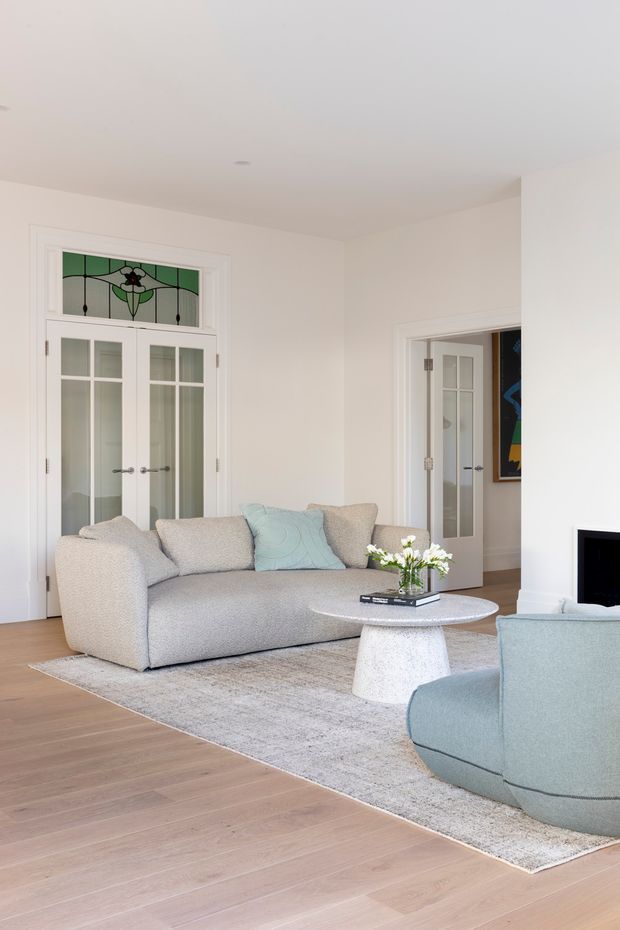 Bright, refined living room with a soft neutral palette. The room features a large light-grey curved sofa, pale blue accent chair and cushion, textured light rug, round stone-look coffee table, pale timber flooring, white walls and white-framed glazed internal doors. A decorative stained-glass transom window adds a traditional architectural detail.