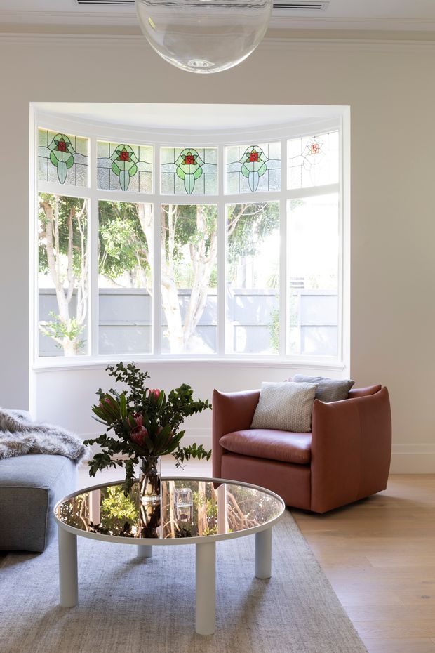 Bright, refined living room with a large bay window featuring decorative stained-glass transom panels. The room has light neutral walls, timber flooring, a grey sofa, a sculptural brown leather armchair, a round reflective coffee table, and a pale area rug. Abundant natural light and greenery outside create an airy, inviting atmosphere.
