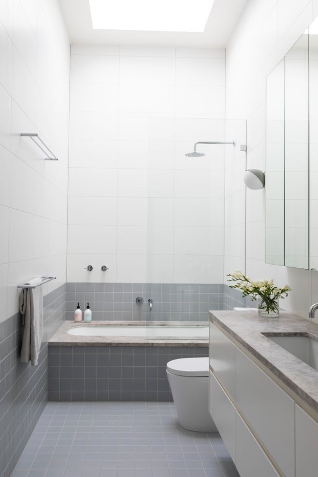 A bright, compact contemporary bathroom with a freestanding bathtub set into a tiled surround, wall-mounted shower, toilet, large mirrored cabinet, and a long grey stone vanity with integrated basin. White upper wall tiles contrast with muted grey-blue lower wall and floor tiles, while a large skylight provides abundant natural light.