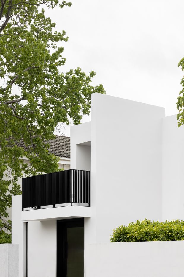 A minimalist white residential facade viewed from outdoors, featuring clean geometric volumes, a recessed entry, and a black metal balcony balustrade. Mature leafy trees, a hedge, and a neighbouring tiled roof frame the contemporary renovation.
