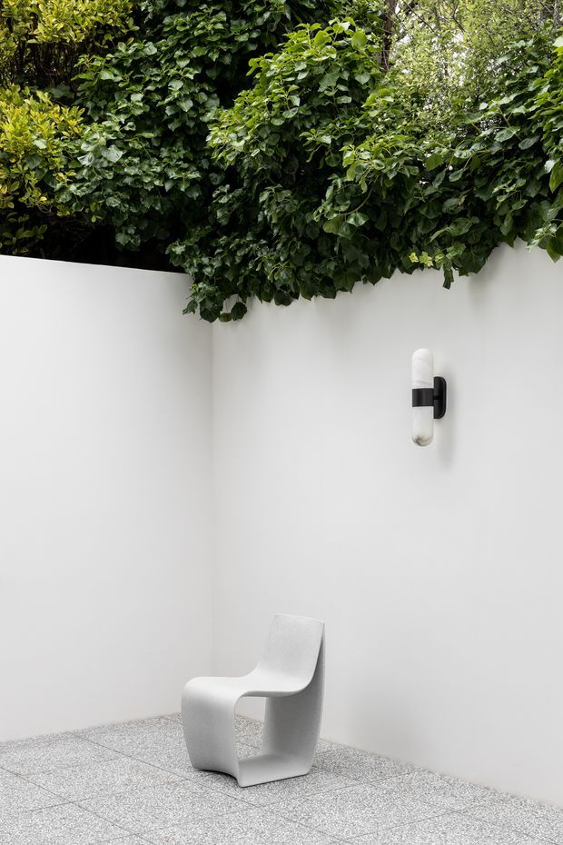 Minimalist courtyard or outdoor architectural nook with smooth white rendered walls, a sculptural white chair, a black-and-white wall sconce, and dense green foliage spilling over the wall edge. The pale terrazzo-like paved floor and strong contrast between white surfaces and greenery create a clean, serene composition.