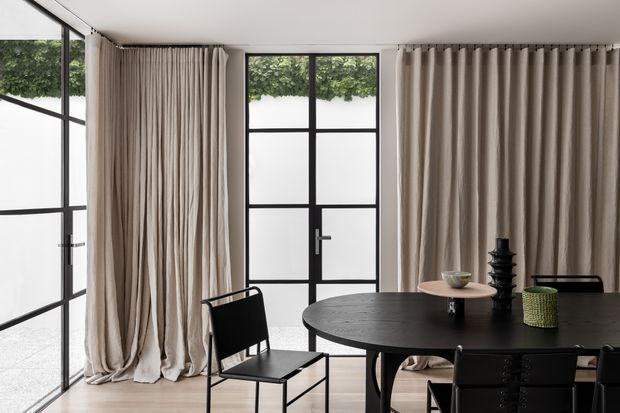A refined dining area with a large round dark dining table and black contemporary chairs, framed by full-height black steel-look glazing and soft beige floor-to-ceiling curtains. The room has pale timber flooring, a light ceiling, and abundant diffused natural light, creating a restrained, elegant atmosphere.