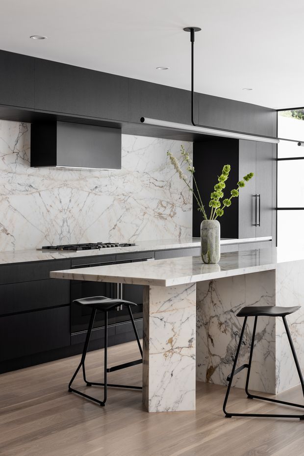 A refined contemporary kitchen with full-height dark cabinetry, a dramatic white veined stone splashback, matching waterfall-edge island, integrated black appliances and slim black pendant lighting. Two black stools sit at the island, while a vase with greenery provides a small natural accent. Light timber flooring and expansive black-framed glazing brighten the space.