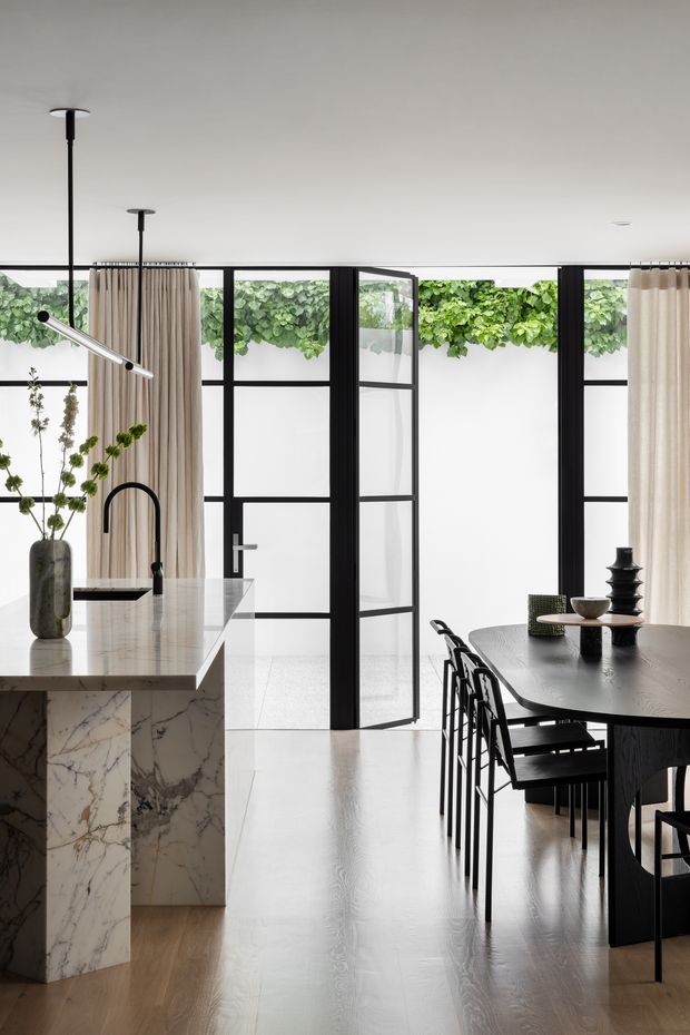 A refined open-plan kitchen and dining area with full-height black-framed glazing and a glazed door opening to lush greenery. The kitchen features a large white marble island with an integrated black undermount sink and curved black tap, pale timber flooring, and neutral full-height curtains. A dark timber dining table with black metal-framed chairs occupies the right side, complemented by sculptural decorative objects and contemporary pendant lighting. The space is bright, calm, and highly finished, with strong indoor-outdoor visual connection.