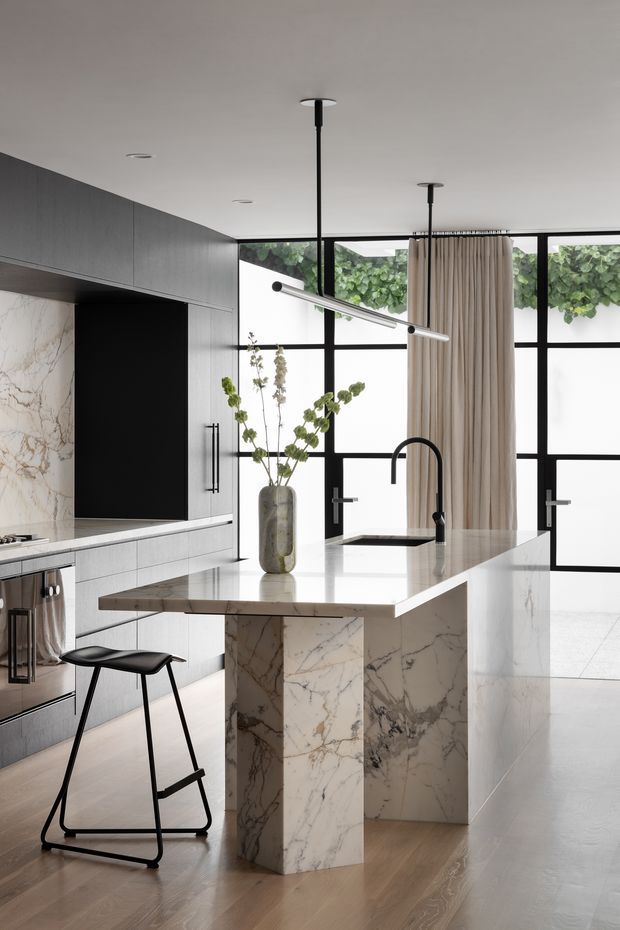 A refined contemporary kitchen with a large marble-clad island, integrated dark cabinetry, a black gooseneck tap, and a slim linear pendant light. Floor-to-ceiling black-framed glazing and a full-height neutral curtain create a bright, open backdrop, while a minimalist black bar stool and sculptural vase add restrained furnishing accents.