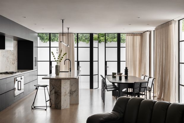 Open-plan contemporary kitchen and dining area with a dark built-in kitchen, marble island, black-framed floor-to-ceiling glazing, sheer curtains, dining table, and a partially visible sofa. The space is bright and refined, with a neutral palette and strong indoor-outdoor connection.
