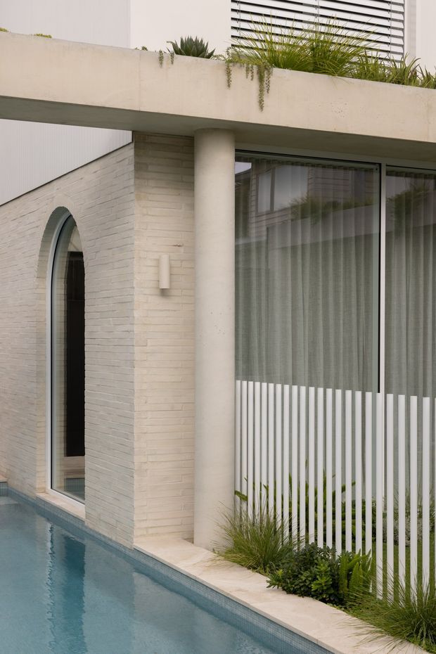 Close-up exterior view of a refined contemporary residence beside a narrow swimming pool. The facade combines pale textured brickwork, a rounded concrete column, a deep projecting terrace or roof slab with planted greenery, and large glazed openings with sheer curtains. A vertical white screen partially conceals the glazing, while an arched doorway and narrow pool edge add architectural detail.