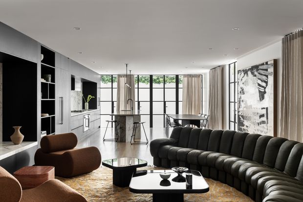 A spacious open-plan contemporary living area with a kitchen and island at the rear. The room features a large curved dark modular sofa, sculptural brown lounge chairs, two black coffee tables, a pale patterned rug, extensive black-framed glazing with sheer curtains, and a large monochrome artwork. The kitchen has dark and light built-in cabinetry, a stone island benchtop, integrated appliances, and pendant lighting. The palette is predominantly monochromatic with warm brown and beige accents.