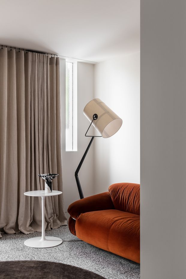 A compact, softly lit living room corner viewed through a doorway or opening. The space features an off-white ceiling and walls, full-height taupe curtains, a narrow vertical window, a rust-orange upholstered sofa, a black angled floor lamp with a beige drum shade, a small white pedestal side table, and a speckled grey carpet. A dark round rug is partially visible in the foreground. The interior has a refined modern, minimalist composition with warm furnishings and textured textiles.