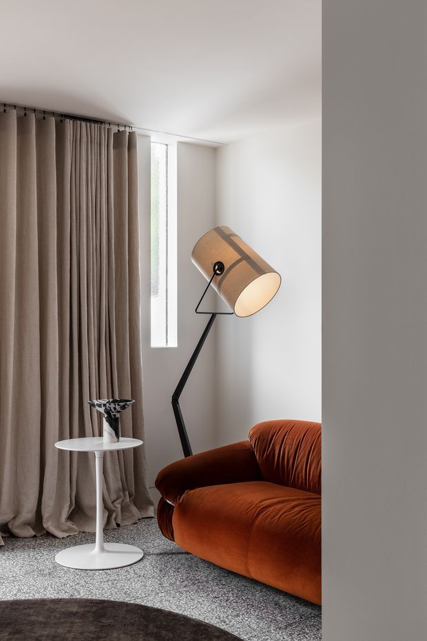 A refined living room corner with a rust-orange upholstered sofa, sculptural black floor lamp, small white pedestal side table, full-height taupe curtains, and a narrow vertical window. The room has white walls and ceiling, a textured grey carpet, and a partially visible dark round rug. The composition is minimalist and softly lit, with a warm, sophisticated palette and mid-century-inspired furnishings.