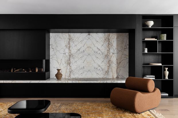 A sophisticated dark-toned living room featuring a full-height marble fireplace surround and hearth, black built-in cabinetry, open shelving with curated decor, and a sculptural rust-brown lounge chair. A patterned area rug sits over a timber floor, creating a refined, dramatic interior.
