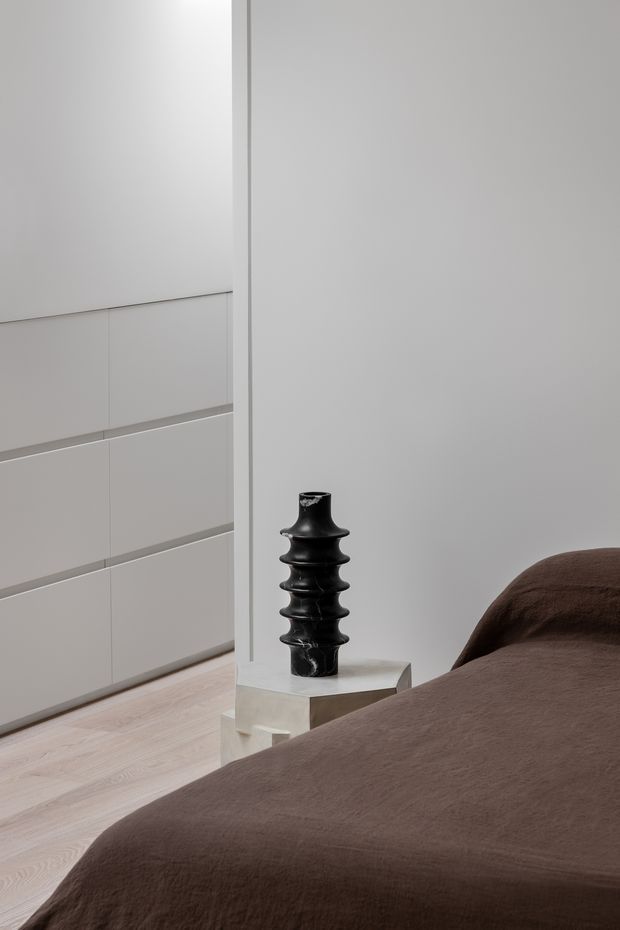 Minimal, softly lit bedroom interior with a low-profile bed dressed in dark brown textured bedding. A black sculptural vase sits on a small pale stone or concrete pedestal beside the bed. The room features white built-in cabinetry with horizontal recessed divisions, pale timber flooring, and plain light-grey walls, creating a restrained contemporary composition.