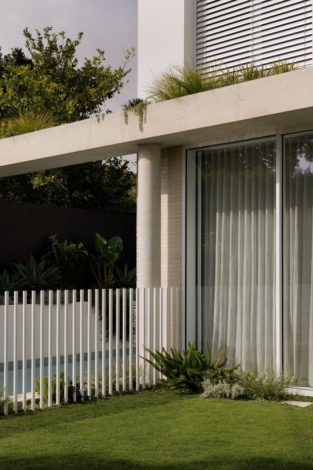 A close architectural exterior view of a contemporary residence, showing a projecting flat concrete terrace or roof slab, round concrete column, pale masonry wall, extensive floor-to-ceiling glazing with sheer curtains, and horizontal external blinds. A white vertical-bar fence encloses a landscaped garden with lawn and tropical planting, with a glimpse of a pool behind the fence.