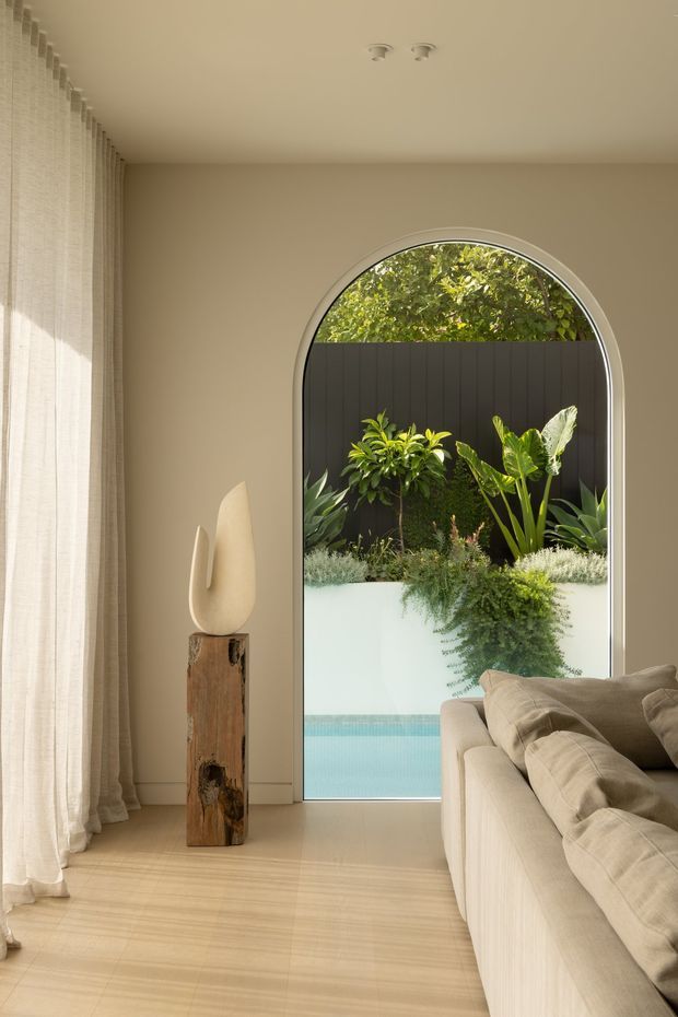 Warm, softly lit living room with pale timber flooring, beige walls and full-height sheer curtains. A large arched glazed opening frames a planted courtyard and blue pool beyond, while a neutral upholstered sofa and sculptural artwork on a rustic timber pedestal furnish the room.