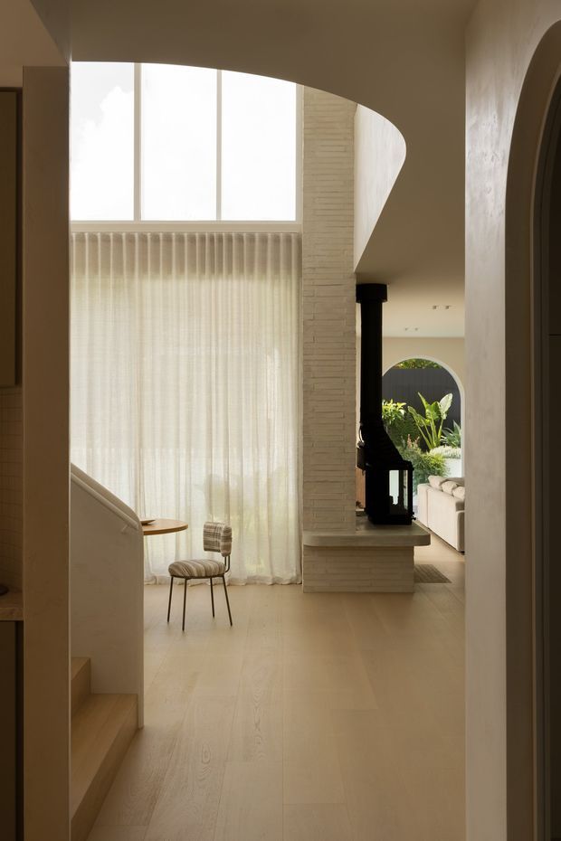 A warm, minimalist interior view centred on a pale timber staircase and open living area. A tall clerestory-style window with sheer curtains floods the space with soft daylight. The room features pale timber flooring, a sculptural curved ceiling and wall forms, a light brick fireplace surround with a black wood-burning stove, and an arched opening leading toward a seating area and greenery outside. A small upholstered chair and side table sit beside the window.