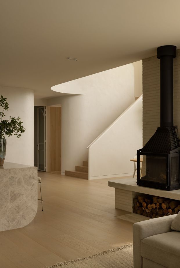 Warm, minimalist open-plan interior featuring a pale timber floor, sculptural curved stone kitchen island, cream plaster walls, a staircase, and a prominent black wood-burning fireplace with integrated log storage. The image is photographed from the living area toward the stair and kitchen zone, with soft neutral furnishings and indirect natural light.