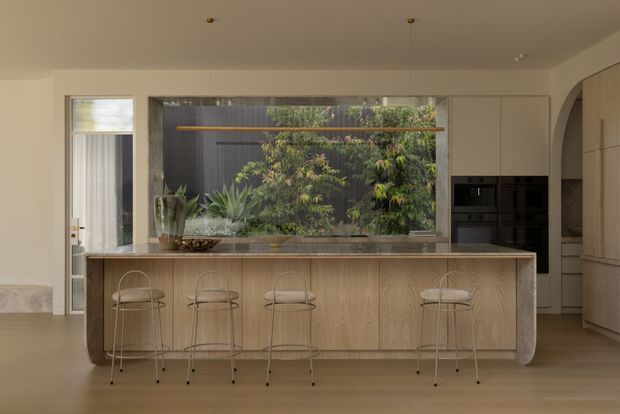 Contemporary kitchen centred on a large timber island with rounded ends and four slim wire-frame bar stools. Integrated dark appliances are set into the cabinetry at right, while a broad glazed opening behind the island looks onto a planted courtyard. Pale walls, timber flooring and restrained detailing create a warm, calm, minimalist composition.