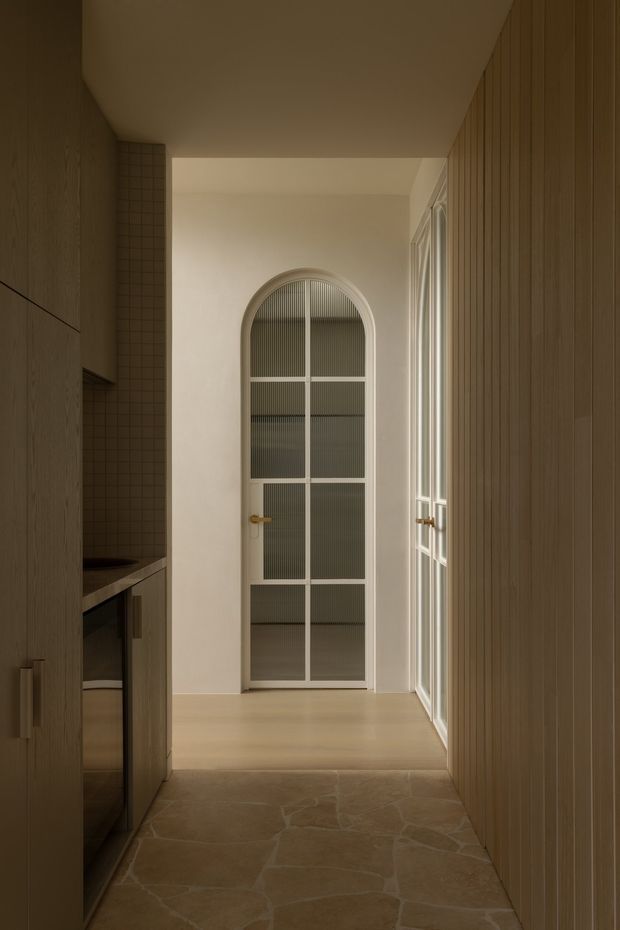 A narrow, warm-toned interior hallway or entry passage leading to an arched glazed door. The space features vertical timber wall panelling on the right, timber cabinetry and a dark benchtop on the left, small square tiled wall detailing, pale timber flooring on the raised rear level, and irregular natural stone or stone-look flooring in the foreground. Tall arched and multi-pane glazing brings soft natural light into the corridor.