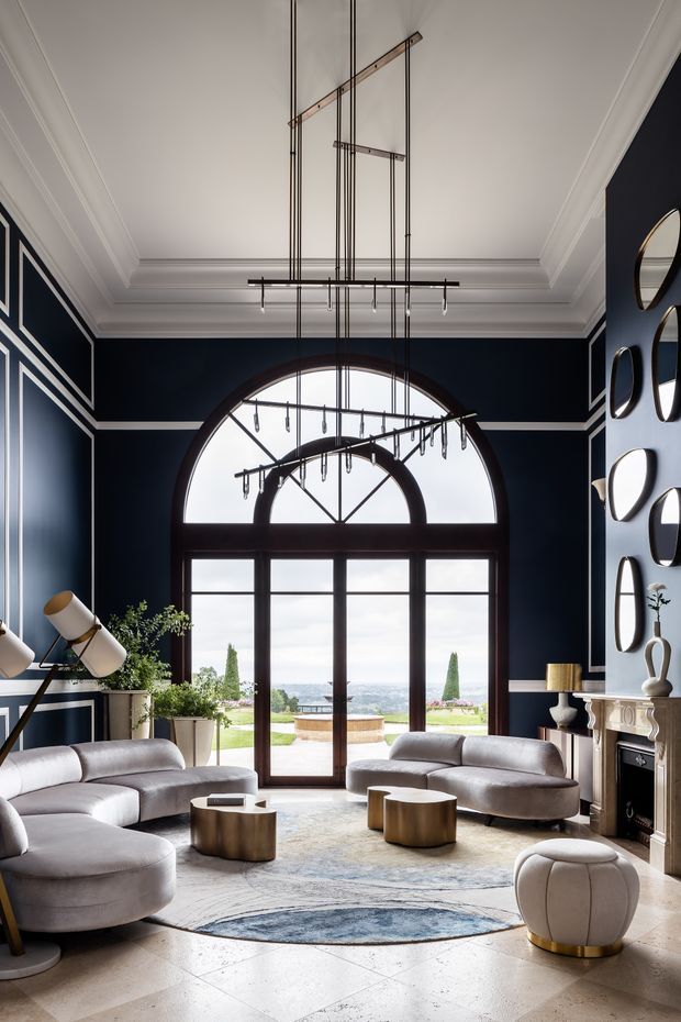 A grand, light-filled formal living room with deep navy walls, crisp white architectural mouldings, a high coffered ceiling, and a dramatic arched window and glazed double doors opening to landscaped grounds and a distant water view. Curved pale sofas and ottomans surround low brushed-gold coffee tables, while a large linear chandelier hangs in the double-height space. Decorative circular mirrors, a classical fireplace mantel, and large potted plants complete the refined interior.