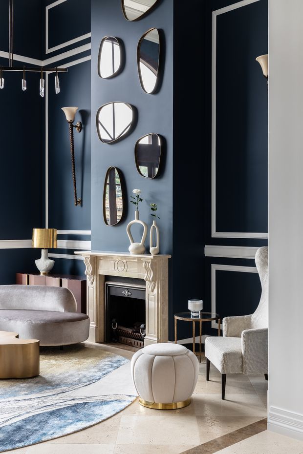 Elegant formal living room with deep navy walls, white decorative panel moulding, a classic stone fireplace, and a curated arrangement of irregular mirrors above the mantel. The room includes sculptural upholstered seating, a rounded ottoman with a brass base, a small side table, a contemporary rug, pendant lights, and refined metallic accents. The overall scheme is dark, luxurious, and polished with a blend of traditional architectural detailing and contemporary furnishings.