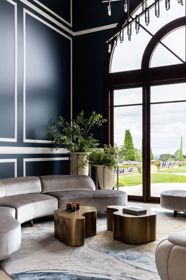 Elegant, high-ceilinged living room with deep navy wall panelling outlined in white trim, a large dark timber-framed arched window and glazed doors opening to landscaped grounds. The room features curved pale upholstered sofas, sculptural bronze-toned nesting coffee tables, potted greenery, a large neutral patterned rug, and a linear chandelier with hanging glass elements. The interior is refined, dramatic, and contemporary with classical detailing.
