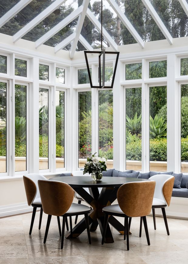 Bright glazed dining sunroom with floor-to-ceiling white-framed windows, a pitched glass roof with exposed white rafters, and a contemporary black geometric pendant light. A dark dining table is surrounded by upholstered brown and white chairs, with a built-in window seat and garden greenery visible outside. The room has pale natural stone flooring and a light, airy atmosphere.