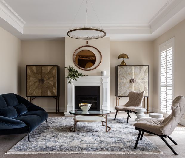 Refined living room with a classic fireplace mantel, round wall mirror, layered artwork, sculptural pendant light, dark blue velvet sofa, cream lounge chairs, glass coffee table, and a large patterned rug. The room features warm neutral walls, white architectural trim, plantation shutters, and balanced contemporary-traditional styling.