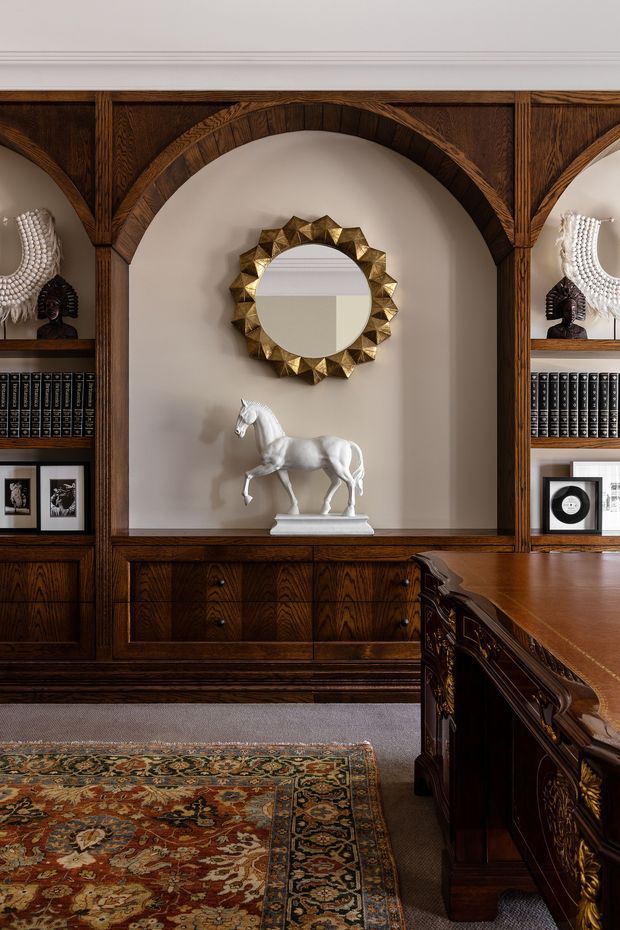 A richly detailed study or home office featuring dark timber built-in cabinetry and open shelving with arched niches, books, framed artwork and decorative objects. A central round sunburst mirror hangs above a white horse sculpture, while an ornate dark timber desk occupies the foreground. The room has a patterned Persian-style rug, muted beige walls, and a refined traditional aesthetic.