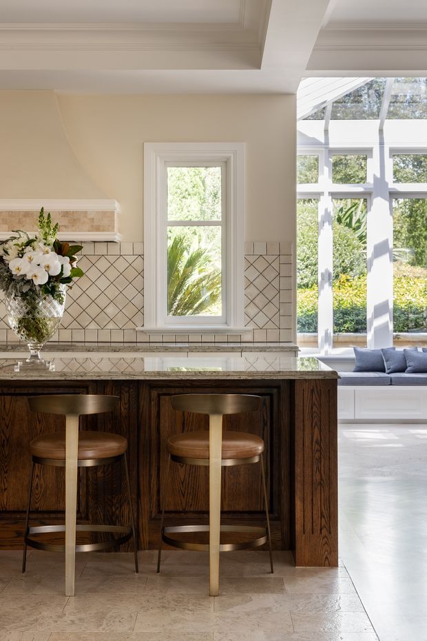 Bright, refined kitchen interior featuring a dark stained timber island with paneled cabinetry, a light stone benchtop, cream tiled splashback with geometric grout lines, and brass-and-leather bar stools. A tall window and adjoining sunroom with extensive white-framed glazing bring in abundant natural light. Decorative ceiling mouldings and a floral arrangement add a traditional, elegant character.