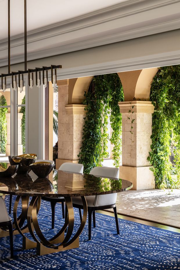 An elegant dining room with a round dark stone dining table, sculptural gold-toned pedestal base, white upholstered chairs, and a dramatic linear pendant light with hanging crystal elements. Ornate painted wall and ceiling mouldings frame large arched openings, which overlook a bright stone terrace with ivy-covered columns and greenery. A deep blue patterned rug adds colour and contrast.
