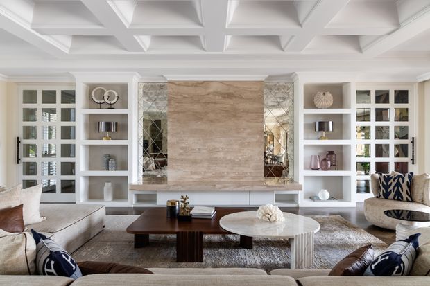 Elegant, symmetrical living room with a large stone feature wall and television recess framed by mirrored panels and white built-in shelving. The room has a coffered ceiling, pale upholstered sofas and armchairs, layered neutral textiles, and dark wood and stone coffee tables, creating a refined, softly luxurious interior.