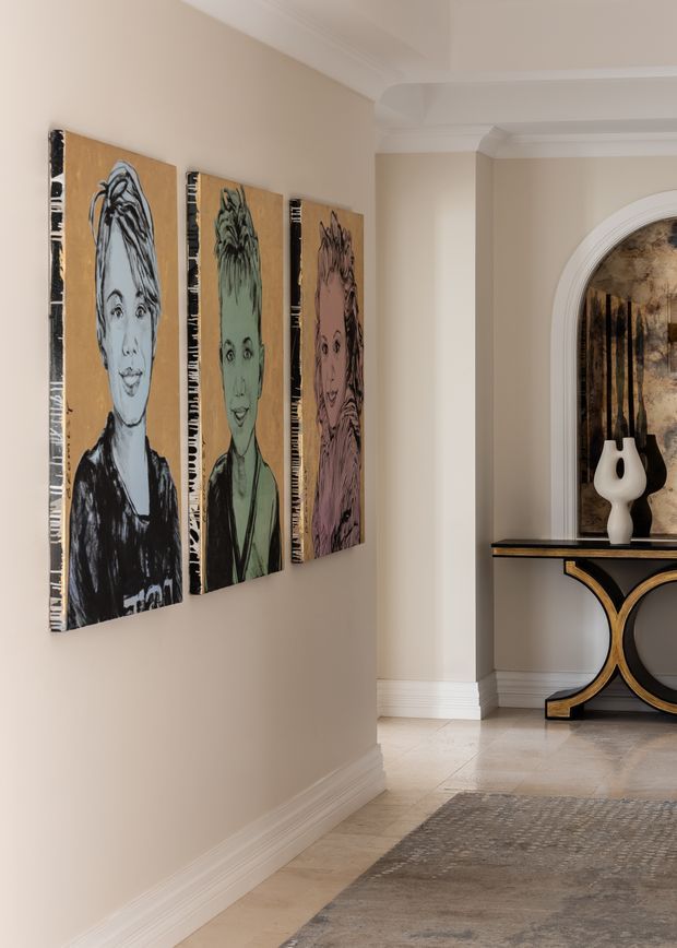 A refined residential hallway or entry passage with warm beige walls, white decorative cornices and skirting, pale stone or tile flooring, and a textured neutral rug. Three vertically arranged portrait artworks line the left wall. At the end, a black console table with curved gold detailing sits beneath a large arched wall opening or mirror, styled with sculptural black-and-white ceramic vases.