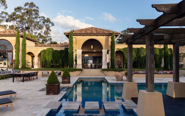 A luxurious Mediterranean-style courtyard featuring a rectangular tiled swimming pool, pale stone paving, clipped hedges, tall cypress trees, terracotta tile roofs, arched colonnades, and a substantial dark timber pergola with outdoor seating. The formal landscape and grand central arch create a refined resort-like atmosphere.