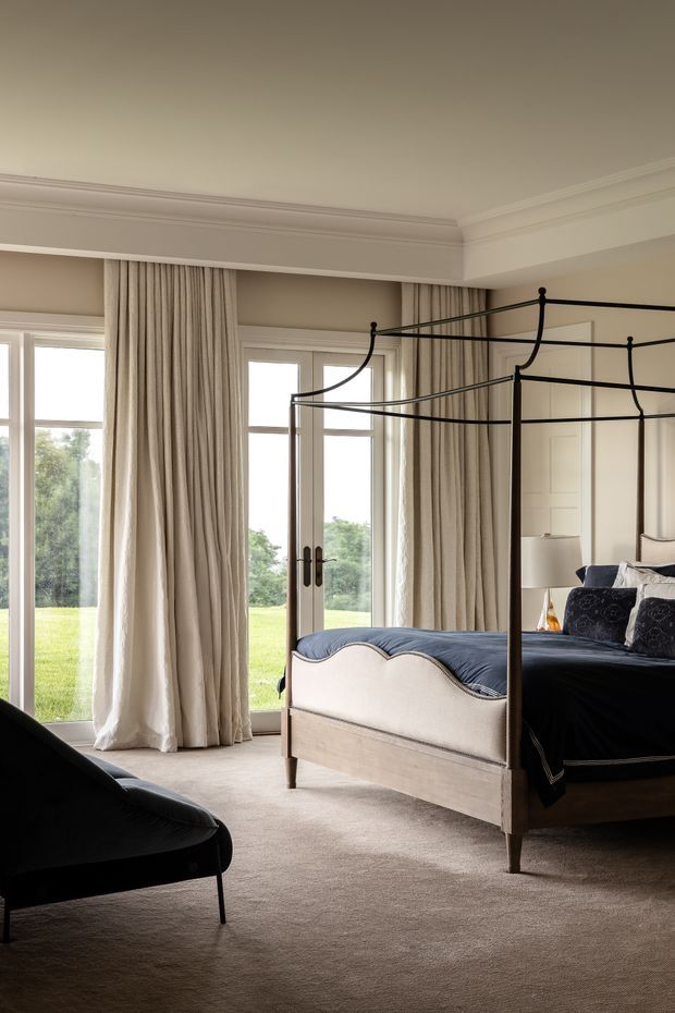 Elegant traditional bedroom with a black metal four-poster bed, upholstered scalloped footboard, dark bedding, patterned cushions, floor-to-ceiling cream curtains, French doors, pale carpet, and detailed ceiling cornices. The room has abundant natural light and views onto a green lawn.