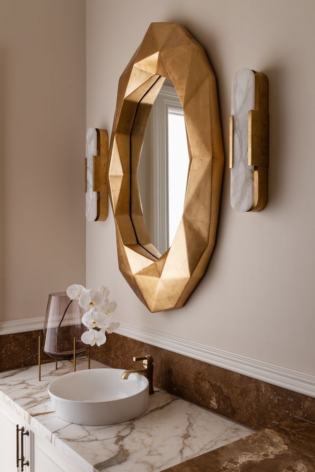 A refined bathroom vanity featuring a white vessel basin on a dramatic white marble countertop, dark brown stone backsplash and flooring, and a sculptural faceted timber-framed mirror. Decorative wall sconces, a glass vase with white orchids, and warm neutral tones create a luxurious, artful atmosphere.