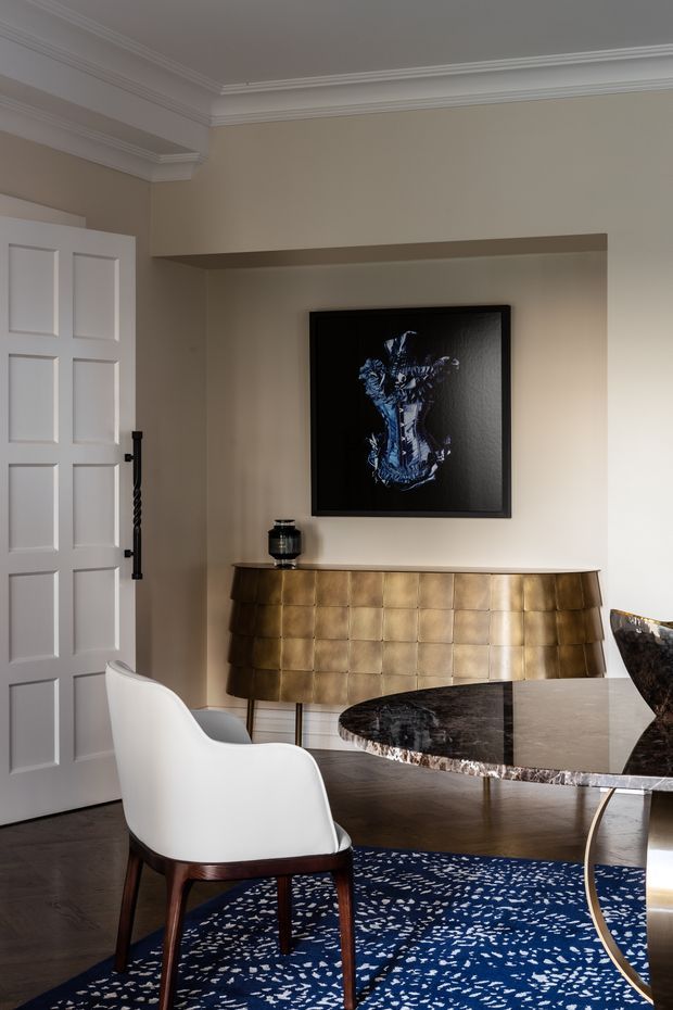 Elegant dining area with a refined, traditional architectural shell. The room features warm taupe walls, ornate white crown moulding, a white paneled door, dark timber flooring, a sculptural white dining chair, a dark stone dining table with a highly irregular natural edge, a blue patterned rug, and a striking brass-toned tiled sideboard beneath framed artwork.