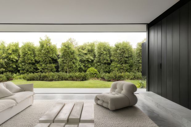 A serene contemporary living room with a broad floor-to-ceiling opening overlooking a manicured lawn and dense hedging. The room features a pale upholstered sofa, sculptural rounded lounge chair, low stepped rectangular coffee table, pale textured rug, light flooring, a smooth white ceiling, and a dramatic full-height dark vertical-panel feature wall with concealed or minimal detailing.
