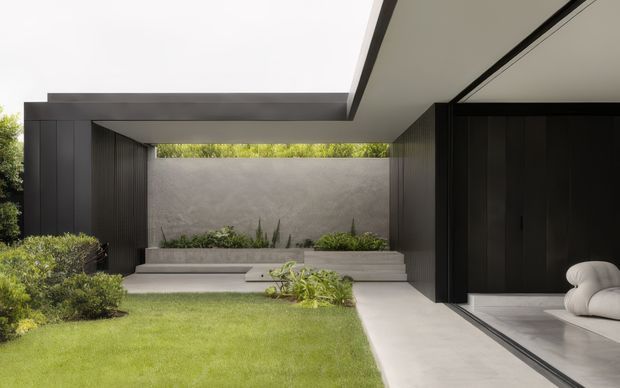 A refined modern courtyard viewed from a covered outdoor passage, with dark vertical-clad walls, a broad projecting flat roof, pale concrete paving, raised planter beds, lush low landscaping and a manicured lawn. A large glazed opening on the right reveals a minimalist interior seating area with a white sculptural sofa. The composition emphasizes strong horizontal rooflines, an enclosed courtyard and indoor-outdoor connection.