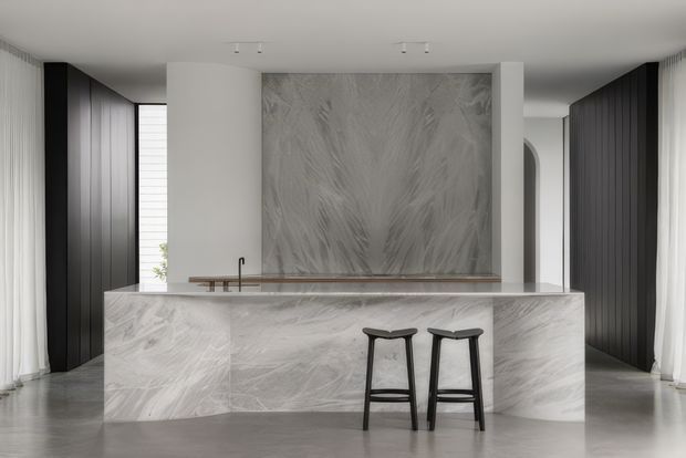 A contemporary minimalist kitchen dominated by a large pale grey veined stone island and matching full-height feature backsplash. Two dark timber or black stools sit at the island, while dark vertical cabinetry lines both side walls. Sheer curtains, pale walls, polished flooring, and restrained recessed lighting create a refined monochromatic interior.