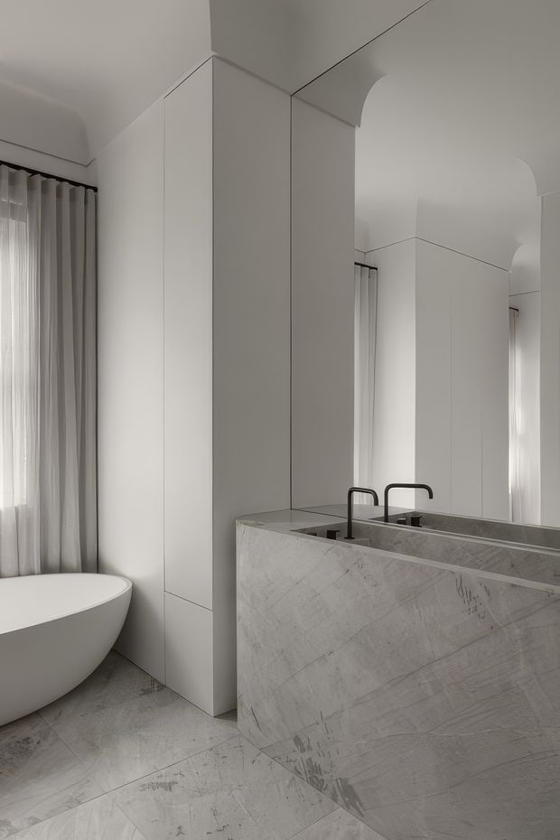 Minimalist monochromatic bathroom with a freestanding oval bathtub beside sheer curtained glazing, a large stone-clad vanity with twin black taps, full-height mirrors, concealed cabinetry, and softly curved arched ceiling transitions. The room uses pale grey veined stone throughout with white surfaces and restrained detailing.