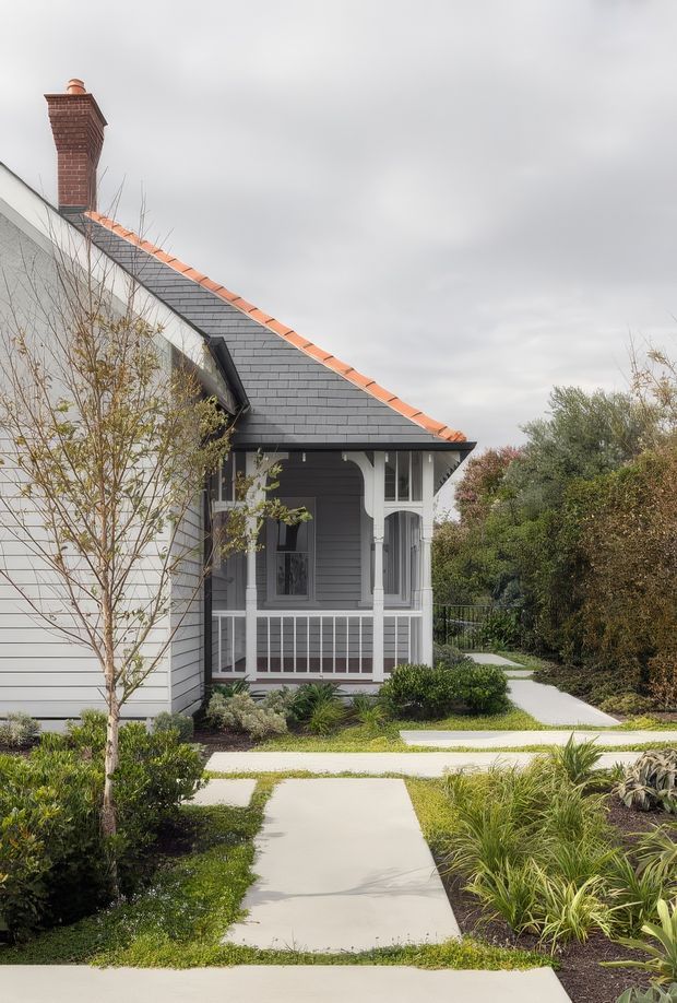 Exterior view of a traditional gabled house with dark grey shingle roofing, pale weatherboard cladding, a red brick chimney, and a white-trimmed enclosed front porch. Pale concrete garden paths pass through a landscaped front garden with shrubs and ornamental grasses under an overcast sky.
