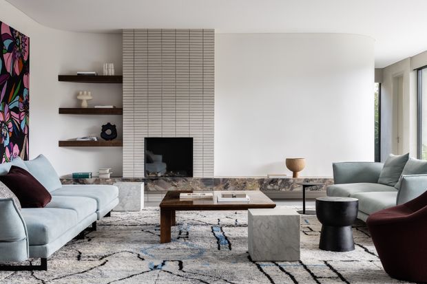 A refined contemporary living room with a sculptural curved white fireplace wall, vertically tiled fireplace surround, dark timber floating shelves, marble hearth, and large windows. Pale sofas, a dark wood coffee table, marble side table, sculptural black side table, and a patterned area rug create a sophisticated neutral scheme with colourful artwork as an accent.
